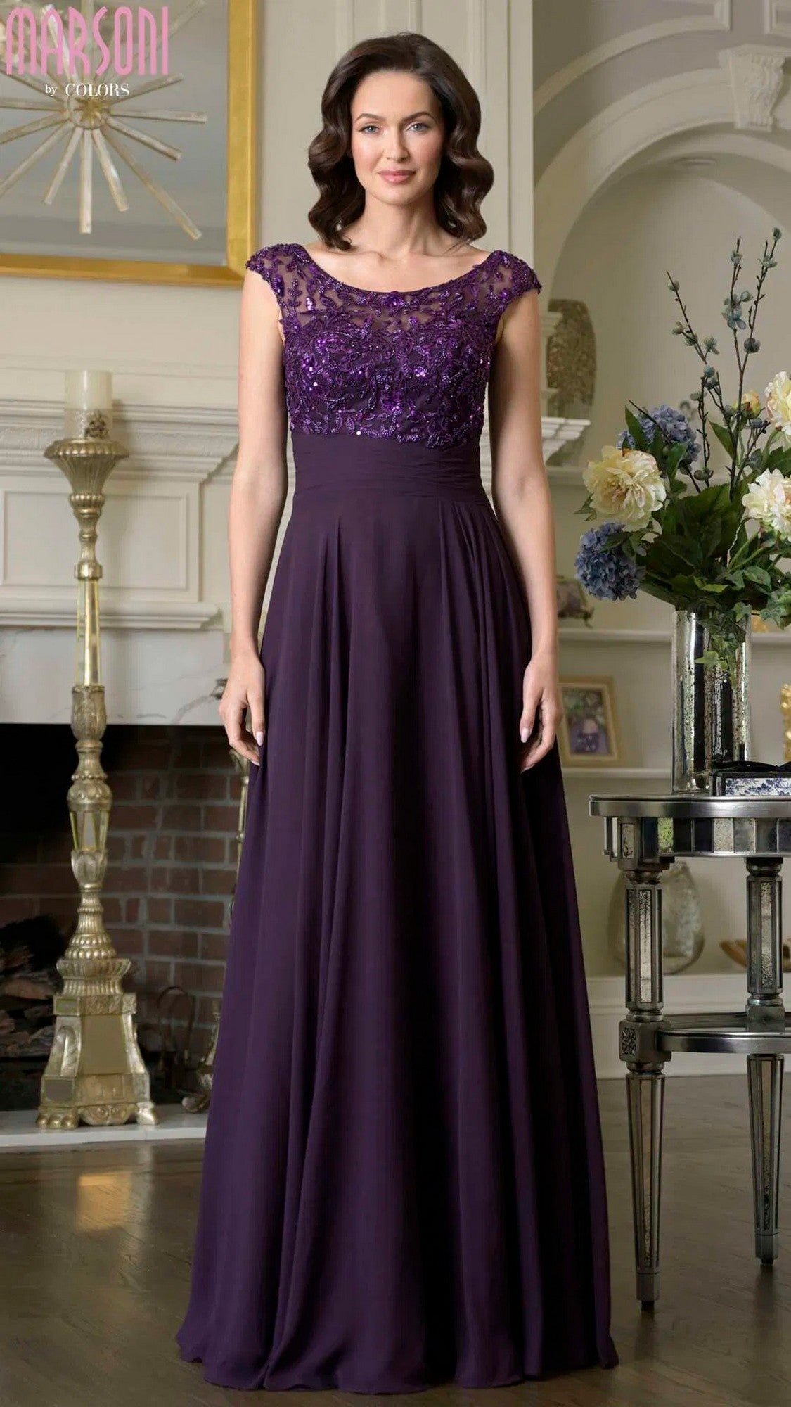 A woman stands indoors wearing the Marsoni M335 long formal dress in eggplant, featuring a floor-length chiffon a-line skirt, fitted sequined bodice, and elegant cap sleeves.